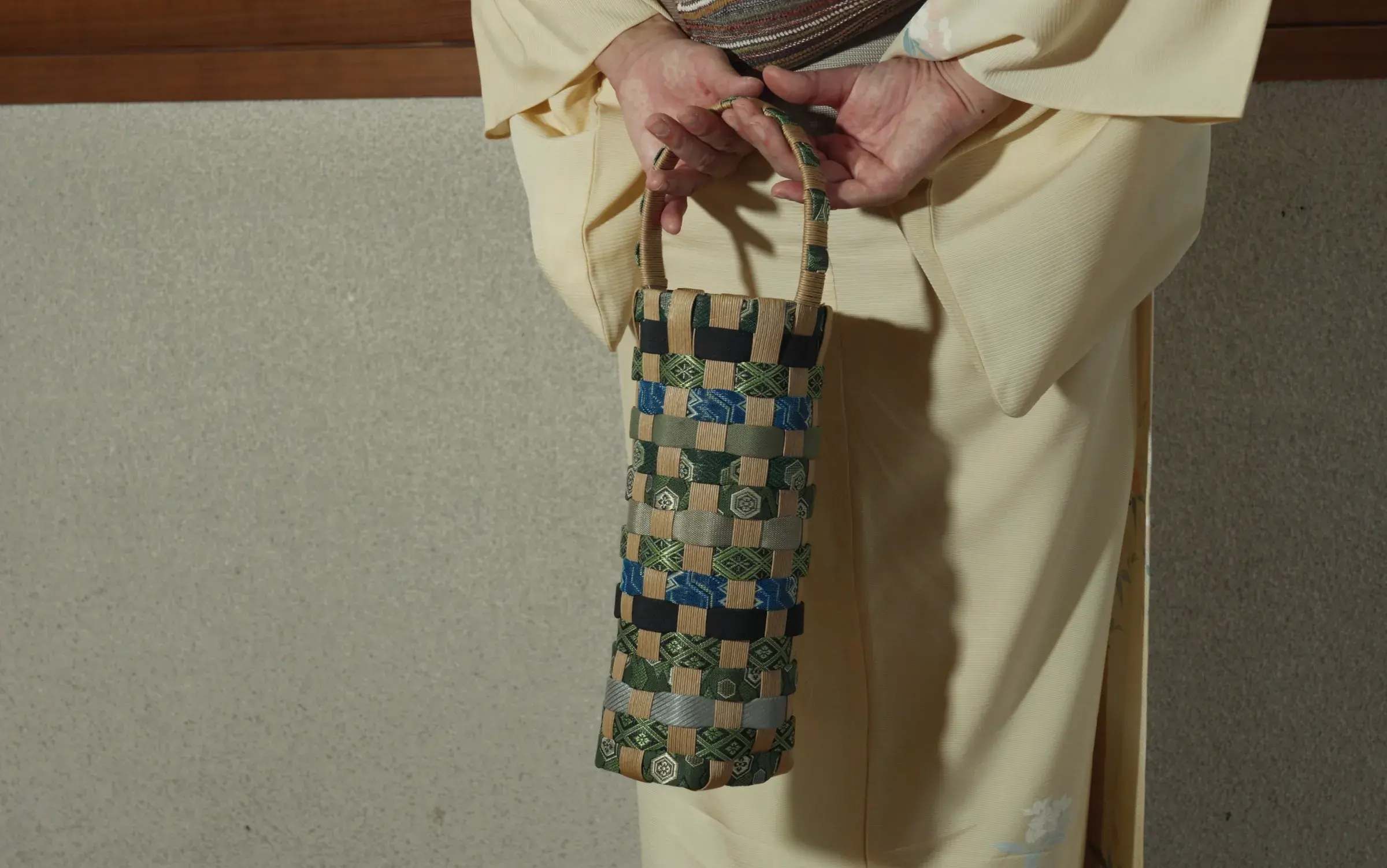 Hands held behind the back, a woven tatami-beri bottle bag hanging from them